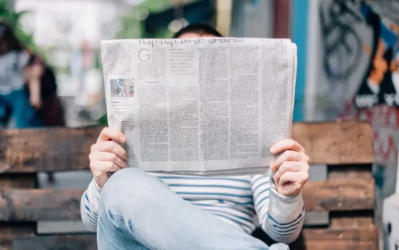 A person sits on a bench while reading a newspaper and looking for information about the Corona crisis. 