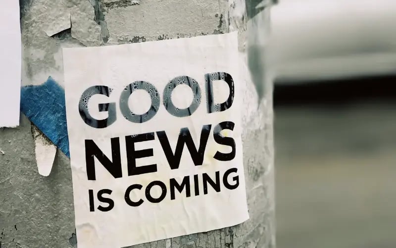The words "Good News Is Coming" is printed in black on a white piece of paper and hangs on a pole. 