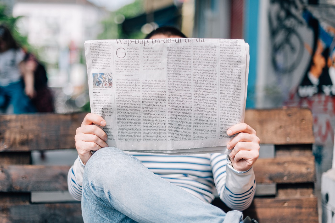 A person sits on a bench while reading a newspaper and looking for information about the Corona crisis.