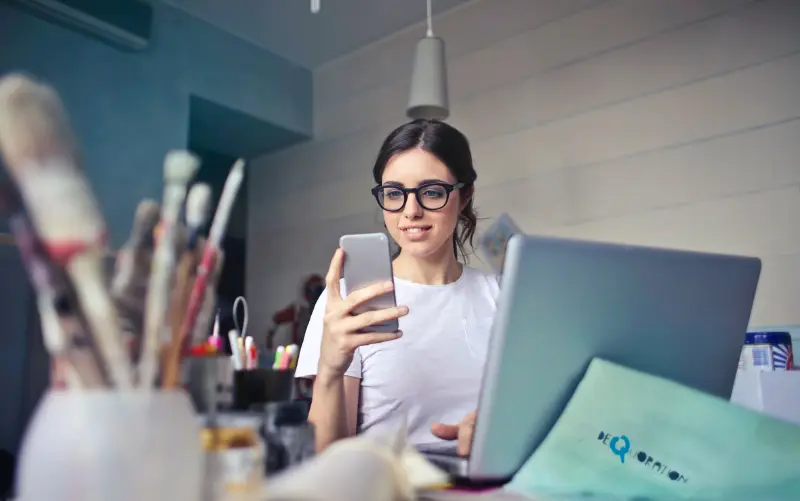 Eine Frau schaut auf ihr Handy, während sie vor einem Laptop sitzt. Vielleicht sucht sie in einem Liveblog nach einem Live-Update. 