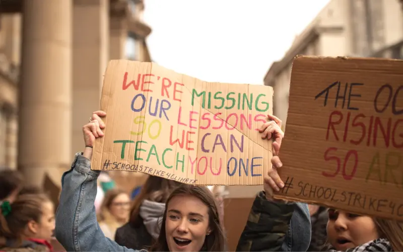 A girl holds a cardboard sign above her head in a crowd of students. Her sign says: WE'RE MISSING OUR LESSONS SO WE CAN TEACH YOU ONE. 