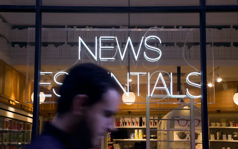 Ein Mann geht an einem Fenster vorbei, an dem eine weiße Leuchtreklame mit der Aufschrift News Essentials angebracht ist. 