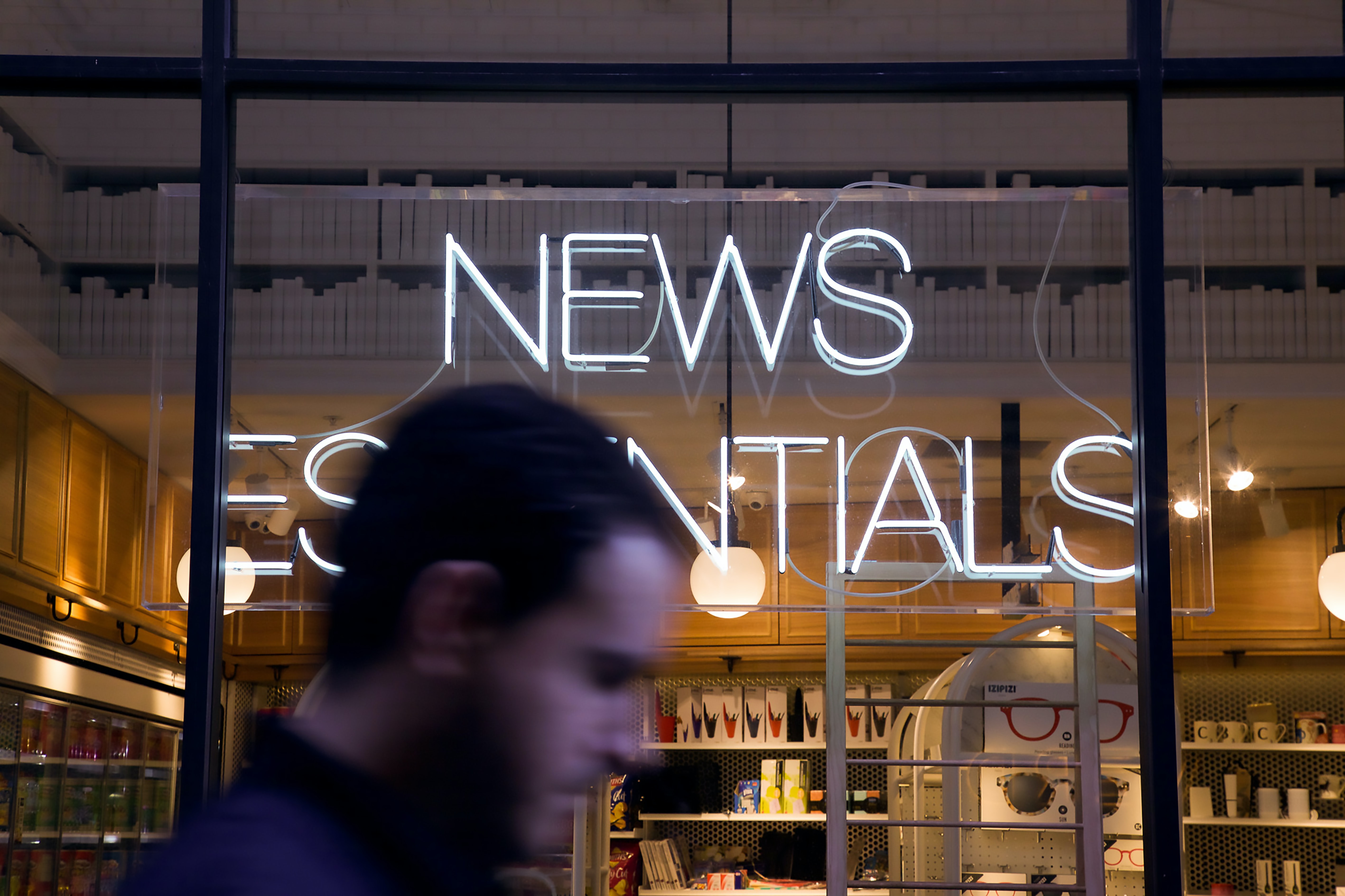 Man walks past a window which has a white neon sign which is made up of  the words News Essentials 